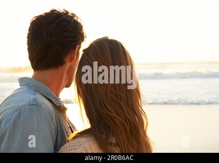 Romance authentique. Un jeune couple s'embrassant et regardant le soleil se coucher sur la plage. Banque D'Images
