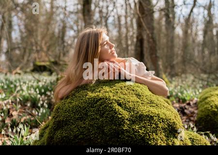 Snowdrops galanthus blond. Une fille en robe blanche s'est couché sur une pierre dans la mousse dans un pré avec des gouttes de neige dans une forêt de printemps Banque D'Images