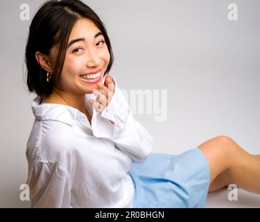 Smiling Young Asian Woman assis sur fond blanc jupe courte manches longues | vue latérale | regarder l'appareil photo Banque D'Images