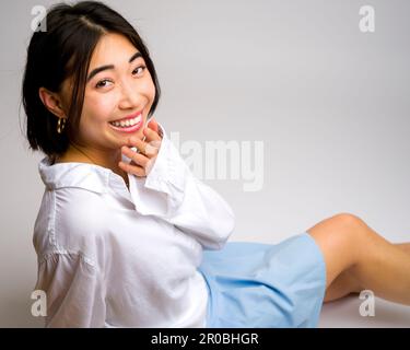 Smiling Young Asian Woman assis sur fond blanc jupe courte manches longues | vue latérale | regarder l'appareil photo Banque D'Images