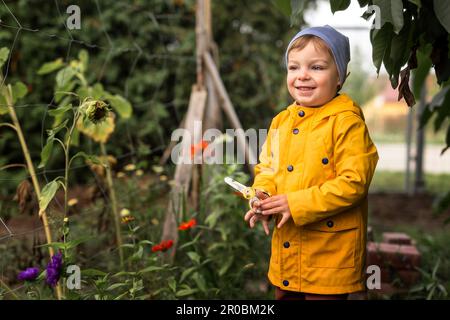 Garçon en imperméable jaune tenant des ciseaux dans le jardin et souriant Banque D'Images