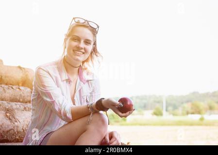 Jeune femme assise sur une pile de bois et tenant une pomme, Bavière, Allemagne Banque D'Images