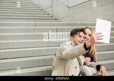Jeune couple assis dans les escaliers et prenant un selfie avec une tablette numérique, Munich, Bavière, Allemagne Banque D'Images