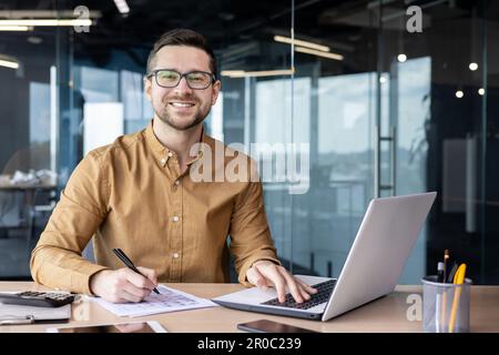 Portrait d'un jeune homme d'affaires en chemise, homme souriant et regardant la caméra sur le lieu de travail à l'intérieur du bureau, comptable avec calculatrice derrière le travail papier signant les contrats et les rapports financiers. Banque D'Images