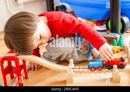 Gros plan d'un enfant de 2 ans de race blanche, garçon, qui se croise sur un plancher dans une pièce, se concentre sur pousser le long d'un moteur de train sur une voie de chemin de fer en bois. Banque D'Images