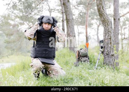 Un homme dans un uniforme militaire et un gilet à l'épreuve des balles travaille dans la forêt pour déminer le territoire. Un homme met un casque de protection avant de commencer le travail Banque D'Images