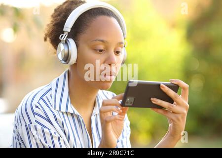 Femme noire sérieuse portant un casque pour regarder des vidéos au téléphone dans un parc Banque D'Images