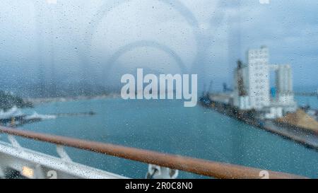 bâtiments sur un quai portuaire flou vus d'un bateau de croisière à travers une fenêtre vitrée recouverte de gouttes de pluie Banque D'Images