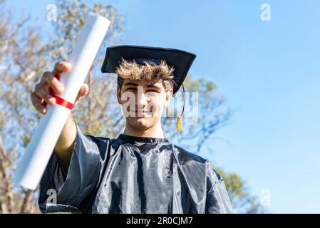 Heureux diplômé jeune homme portant une robe de baccalauréat et un mortarboard noir et montrant son diplôme Banque D'Images