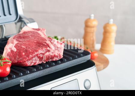 Steaks de première qualité crus sur le gril électrique. Steaks de viande de choix black angus Ribeye sur un grill électrique moderne au romarin, à la tomate cerise et au poivre. Cuisine pro Banque D'Images