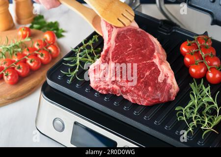 Steaks de première qualité crus sur le gril électrique. Chef à la main tenant le steak Ribeye sur un grill électrique moderne avec romarin, tomates cerises et poivre. Procédure de cuisson Banque D'Images