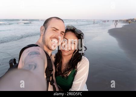 Un jeune couple hispanique et caucasien prend un selfie sur la plage pendant le coucher du soleil Banque D'Images