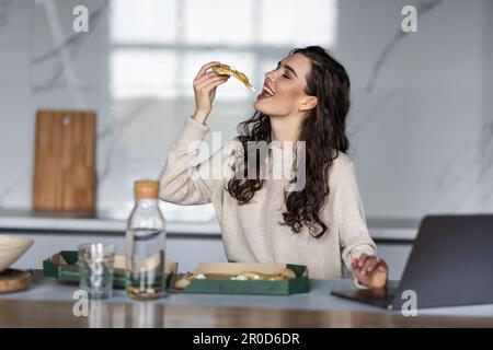 Belle jeune femme avec ordinateur portable manger une pizza savoureuse dans la cuisine Banque D'Images