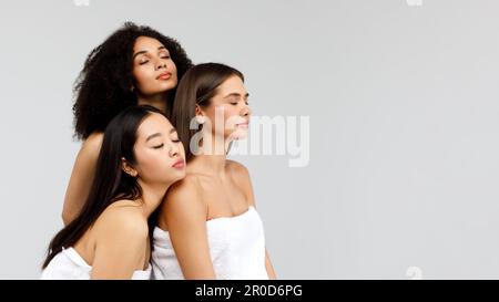 Diversité et beauté des jeunes. Trois femmes mixtes se posant ensemble et regardant de côté à l'espace libre sur fond gris Banque D'Images