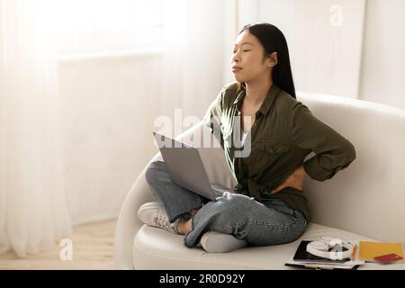 Femme asiatique fatiguée indépendante assise sur un canapé avec un ordinateur portable Banque D'Images