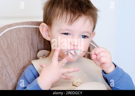 Un enfant heureux mange du porridge avec une cuillère tout en étant assis sur une chaise haute. Bébé dans un bavoir mange du porridge de flocons d'avoine lui-même. Enfant âgé d'environ deux ans (un an) Banque D'Images