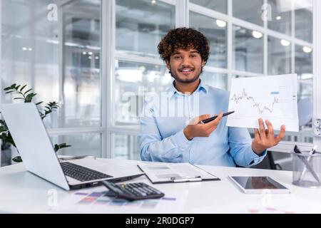 Portrait de jeunes bons documents financiers à l'intérieur du bureau, homme souriant et regardant la caméra montre graphique avec la dynamique positive des bénéfices économiques, homme d'affaires au travail avec ordinateur portable. Banque D'Images