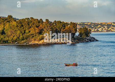 Un petit bateau de pêche retourne au port après une sortie à l'aube. En arrière-plan la ville de Messine. Sicile, Italie, Europe Banque D'Images