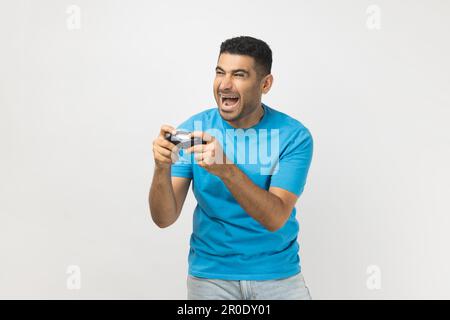 Portrait d'un gamer très heureux et enthousiaste portant un T-shirt bleu debout jouant à des jeux vidéo, tenant le joystick dans les mains. Prise de vue en studio isolée sur fond gris. Banque D'Images