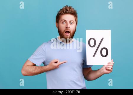 Portrait de l'homme barbu tenant du papier avec l'inscription du signe de pourcentage, regardant la caméra avec la bouche ouverte et l'expression choquée. Studio d'intérieur isolé sur fond bleu. Banque D'Images