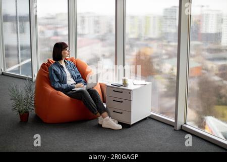 Au-dessus de la vue de la jolie dame aux cheveux foncés en verre, vue sur la fenêtre pleine hauteur avec un panorama étendu sur le centre-ville. Belle femme qui se repose tout en tenant un ordinateur portable sur les genoux. Banque D'Images