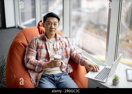 Portrait d'un Indien positif vêtu de façon décontractée, assis sur un pouf de sac de haricot orange et souriant à l'appareil photo dans un bureau confortable. Travailleur souriant tenant une tasse de café aromatique et tapant sur un ordinateur portable. Banque D'Images