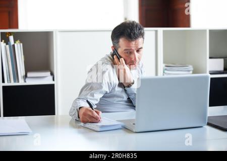 Vérifier les numéros par téléphone... un homme d'affaires épuisé qui parle au téléphone tout en étant assis devant son ordinateur portable et en prenant des notes. Banque D'Images