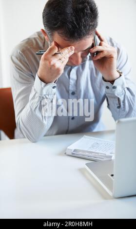 Ces chiffres ne peuvent pas être justes... un homme d'affaires épuisé qui parle au téléphone tout en étant assis devant son ordinateur portable à son bureau. Banque D'Images