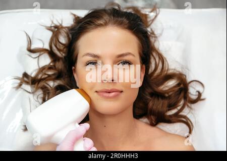 Jeune femme caucasienne positive couché dans un salon de beauté pendant la procédure pour enlever les cheveux indésirables en utilisant l'enlèvement de cheveux au laser. Épilation au laser sur le visage. Procédures du salon Banque D'Images