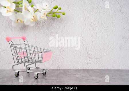 Vide jouet métal supermarché panier sur fond de ciment ancien avec fleur blanche d'orchidée. Banque D'Images