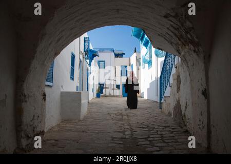 La femme arabe marche à travers l'arcade le long du chemin des bâtiments avec des fenêtres bleues et des murs blancs dans le village artisanal, Sidi Bou Said, Tunis, Tunisie Banque D'Images
