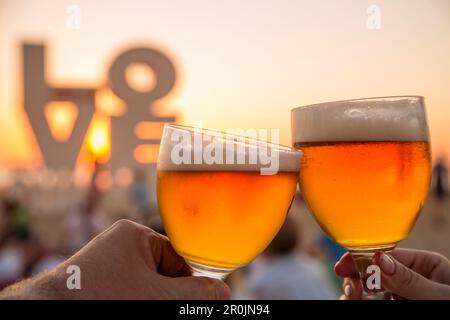 Mains tenir deux verres de bière belge devant la sculpture de l'Amour sur la plage au coucher du soleil, Ostende, Flandre, région flamande, Belgique Banque D'Images
