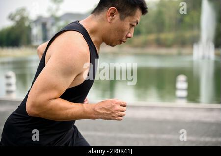 Vue latérale d'un asiatique mûr déterminé et concentré dans des vêtements de sport courant dans un parc public le matin. Banque D'Images