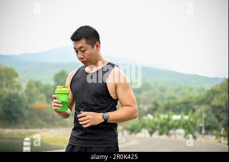 Un homme asiatique mûr et assoiffé de sportswear boit de l'eau dans une bouteille après avoir roulé dans un parc public. Banque D'Images