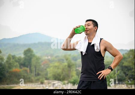 Un homme asiatique mûr et assoiffé de sportswear boit de l'eau dans une bouteille après avoir roulé dans un parc. Concept d'exercice et de loisirs Banque D'Images