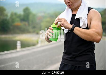 Image rognée, sueur et assoiffé, homme asiatique mûr dans des vêtements de sport, boit de l'eau dans une bouteille après avoir couté dans un parc public. Banque D'Images