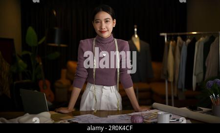 Dessinatrice de mode asiatique millénaire attrayante et réussie ou tailleur debout à son bureau dans son studio. Banque D'Images