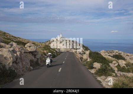 Couple sur un scooter blanc sur la route le long de la péninsule de Cap de Formentor avec phare de Faro de Formentor derrière, Cap de Formentor, Majorque, Iles Baléares Banque D'Images