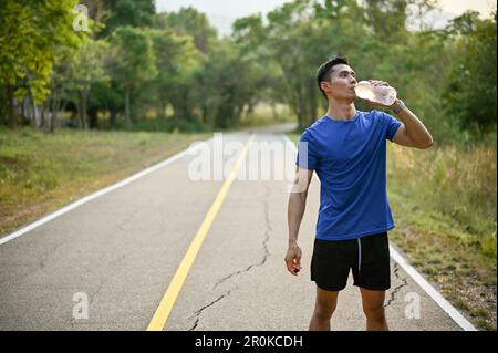 Un homme asiatique assoiffé et épuisé dans des vêtements de sport boit l'eau d'une bouteille tout en courant dans un parc naturel. Concept d'activité et d'exercice en plein air Banque D'Images