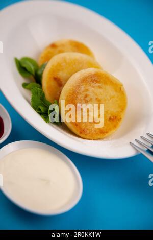 Crêpes rondes au fromage cottage dans une assiette blanche sur fond bleu. Restauration petit déjeuner. Une alimentation saine. Sauce à la crème aigre et menthe. Vue de dessus en gros plan. Dessert. Beignets à la ricotta au fromage caillé. Banque D'Images