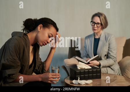 Triste jeune Afro-américaine patiente de conseiller avec un verre d'eau en main décrivant son problème au psychothérapeute en cabinet Banque D'Images