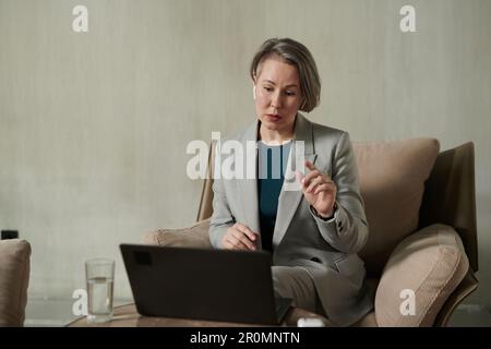 Psychothérapeute mature et confiant dans les écouteurs parlant au patient pendant la session en ligne tout en étant assis dans un fauteuil devant un ordinateur portable au bureau Banque D'Images