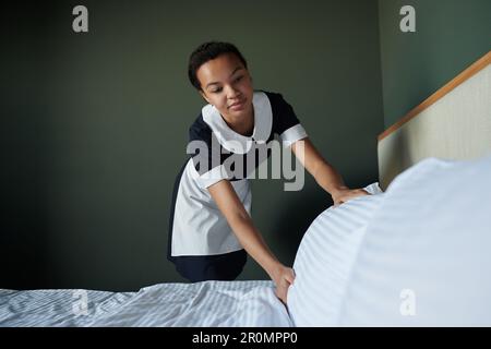 Jeune femme de chambre afro-américaine en uniforme préparant un lit double pour les nouveaux clients de l'hôtel tout en changeant les draps et en redressant l'oreiller Banque D'Images