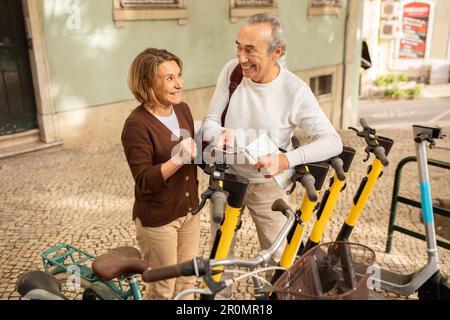 Joyeux couple de touristes senior louant des scooters électriques par téléphone portable à l'extérieur Banque D'Images