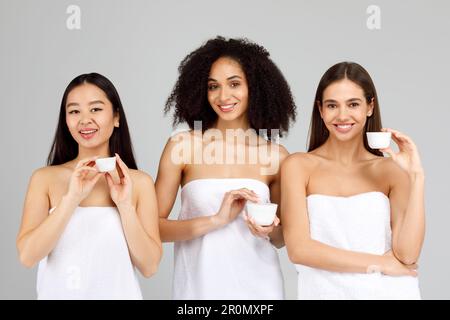 Joyeux diverses dames démontrant produit du visage, nouveaux cosmétiques, posant dans les serviettes de bain sur fond gris Banque D'Images