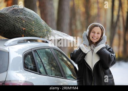 Happy Woman est en train d'écouter de la musique dans des écouteurs, de danser tout en emballant l'arbre de Noël avec le filet et les guirlandes sur un toit de sa voiture, se préparer à un Banque D'Images