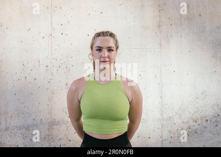 Une jeune femme positive dans les vêtements de sport regardant l'appareil photo tout en se tenant contre un mur de béton avec les mains derrière et souriant pendant l'entraînement de fitness Banque D'Images