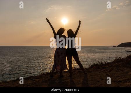 Silhouettes pleines de corps de jeunes amies gaies en tenues d'été levant les bras et profiter du coucher du soleil tout en se tenant près de la mer agitée pendant les vacances Banque D'Images