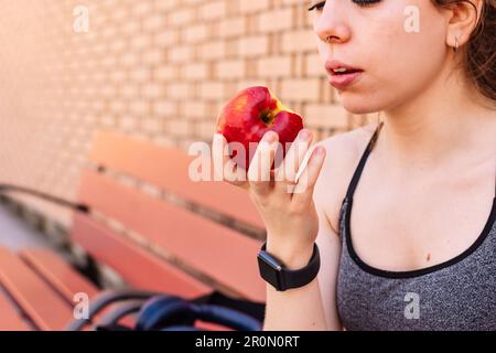 Sportswoman en activité mangeant une délicieuse pomme fraîche tout en étant assis sur le banc pendant la pause fitness sur fond flou Banque D'Images