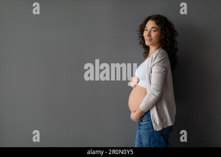 Belle femme enceinte embrassant tendre ventre tout en posant contre le mur de studio gris Banque D'Images